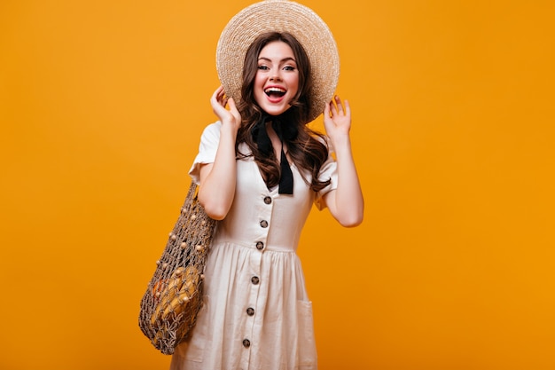 Ragazza carina in abito di cotone in posa con sacchetto di stringa con frutta. La donna in cappello di paglia con fiocco sta ridendo su sfondo arancione.