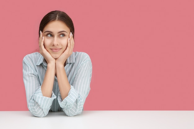 Ragazza carina in camicia a righe elegante seduto al suo posto di lavoro con i gomiti sulla scrivania, cuscino viso sulle mani, distogliere lo sguardo con espressione facciale annoiata o premurosa, pensando a come intrattenere se stessa