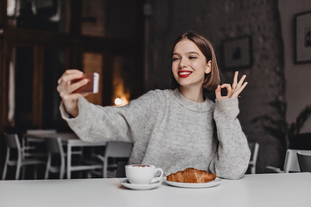 Ragazza carina in maglione grigio prende selfie nella caffetteria e mostra OK. Una donna con un rossetto rosso ha ordinato caffè e croissant.