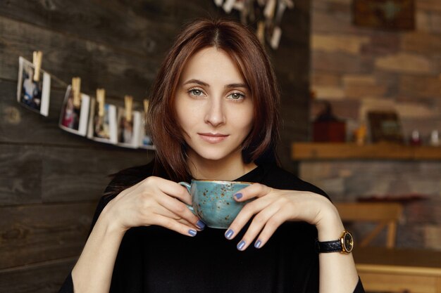 Ragazza carina studentessa con unghie blu che tiene tazza, gustando cappuccino fresco presso la caffetteria