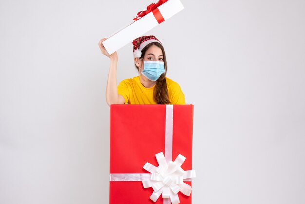 Ragazza carina vista frontale con cappello santa e maschera medica in piedi dietro il grande regalo di Natale