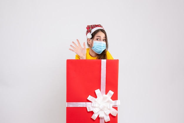 Ragazza carina vista frontale con cappello santa salutando in piedi dietro il grande regalo di Natale