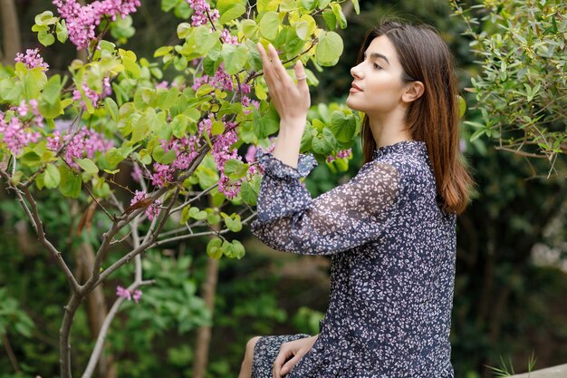 Ragazza castana abbastanza esile che si siede vicino all'albero in fiore che cerca il fiore profumato