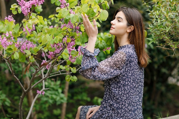 Ragazza castana abbastanza esile che si siede vicino all'albero in fiore che cerca il fiore profumato