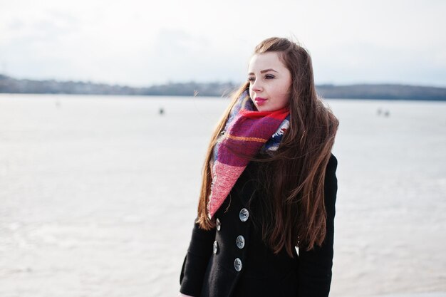 Ragazza casual al cappotto nero sciarpa e cappello contro il fiume ghiacciato in un clima invernale soleggiato