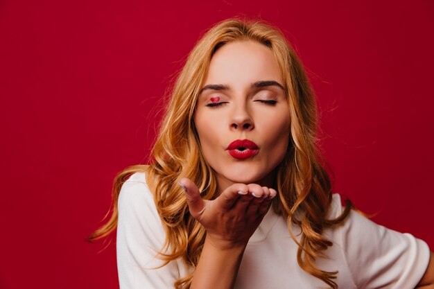 Ragazza caucasica accattivante che esprime amore sulla parete rossa. Affascinante signora bionda che invia un bacio d'aria.