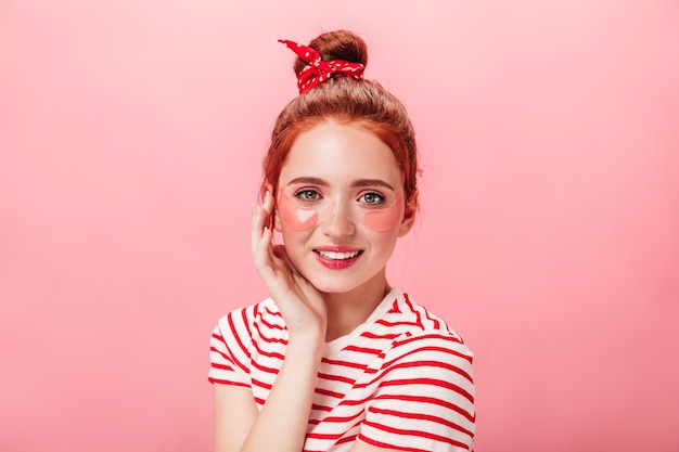 Ragazza caucasica alla moda con bende sugli occhi che guarda l'obbiettivo con un sorriso. Studio shot di zenzero donna in maglietta a righe isolato su sfondo rosa.