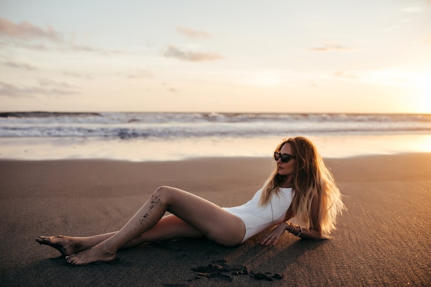 Ragazza caucasica rilassata in costume da bagno bianco che si trova alla spiaggia sabbiosa nella sua vacanza. Foto all'aperto di donna bionda allegra in occhiali da sole a prendere il sole accanto all'oceano e guardando il cielo.