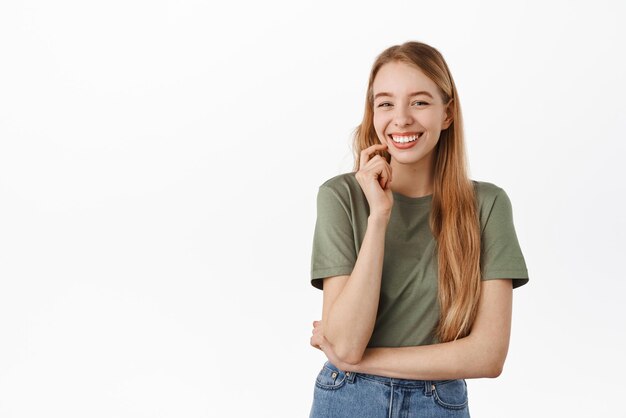 Ragazza caucasica sveglia che ride sorridente con i denti perfetti bianchi che guardano gioiosa alla macchina fotografica come se avesse una conversazione divertente in piedi in maglietta su sfondo bianco