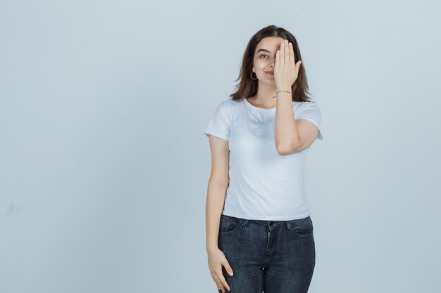 Ragazza che copre gli occhi con la mano in t-shirt, jeans e che sembra felice, vista frontale.