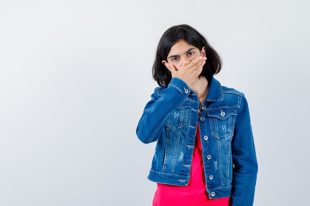 Ragazza che copre la bocca con la mano in maglietta rossa e giacca di jeans e sembra carina. vista frontale.