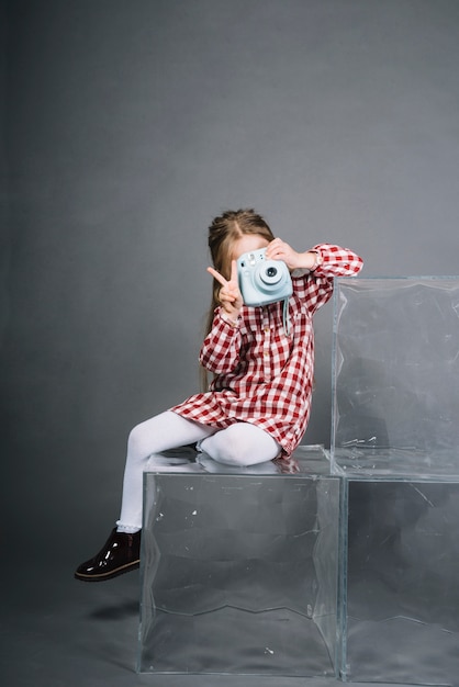 Ragazza che fa clic sulla foto dalla macchina fotografica istantanea che mostra il segno di vittoria contro fondo grigio