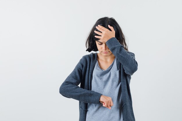 Ragazza che finge di guardare l'orologio mentre mette la mano sulla fronte in t-shirt grigio chiaro e felpa con cappuccio con zip grigio scuro e sembra stressata