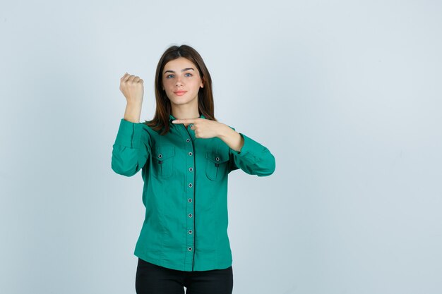 Ragazza che finge di indicare l'orologio al polso in camicetta verde, pantaloni neri e sembra felice, vista frontale.