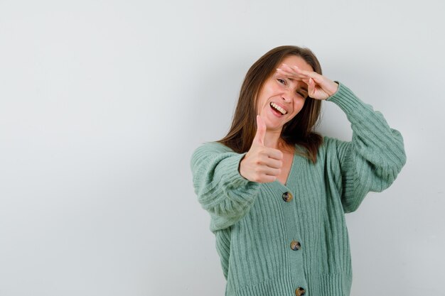 Ragazza che guarda lontano con la mano sopra la testa, mostrando il pollice in maglieria e guardando felice, vista frontale.