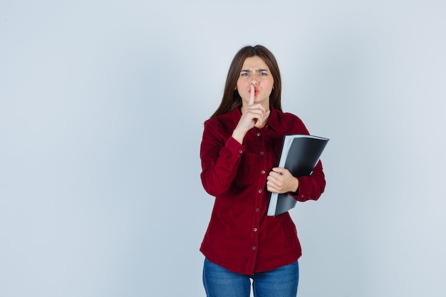 ragazza che mostra gesto di silenzio, tenendo cartella in camicia bordeaux e guardando seria.