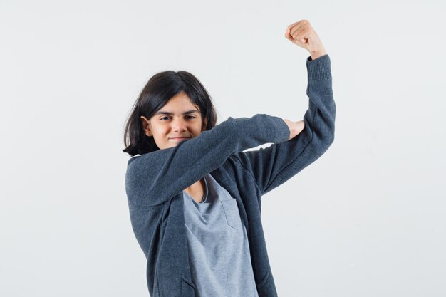 Ragazza che mostra i muscoli in t-shirt grigio chiaro e felpa con cappuccio con zip grigio scuro e sembra carina