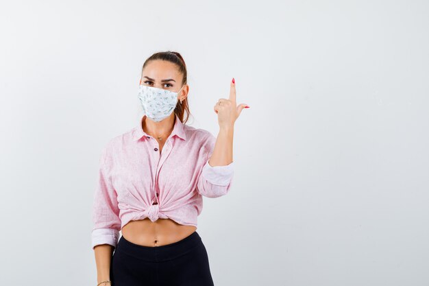 Ragazza che mostra il gesto della pistola in camicetta rosa, pantaloni neri, maschera e sembra seria. vista frontale.