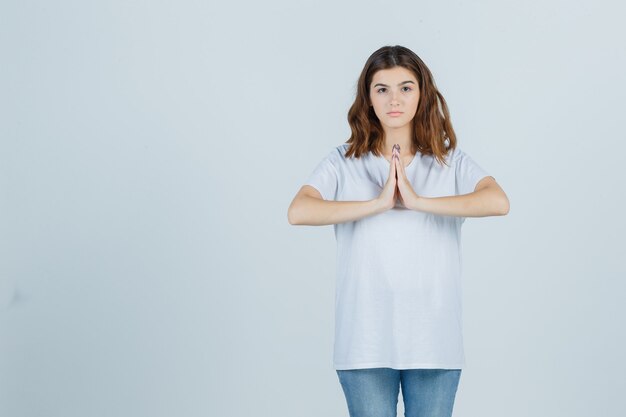 Ragazza che mostra il gesto di namaste in maglietta bianca e che sembra speranzoso, vista frontale.