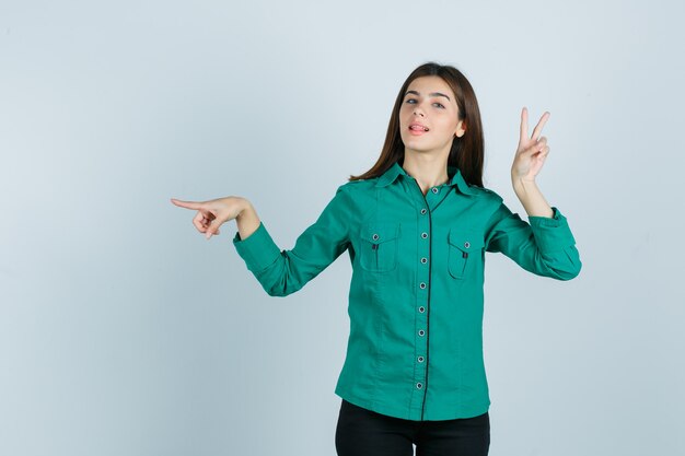 Ragazza che mostra il gesto di pace, puntando a sinistra con il dito indice in camicetta verde, pantaloni neri e guardando fiducioso. vista frontale.