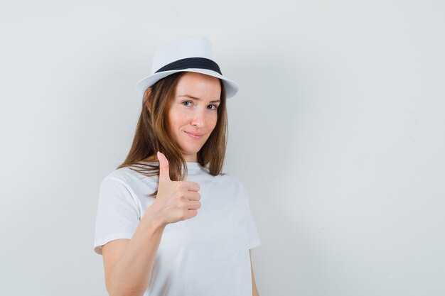 Ragazza che mostra il pollice in su in cappello bianco della maglietta e che sembra fiducioso