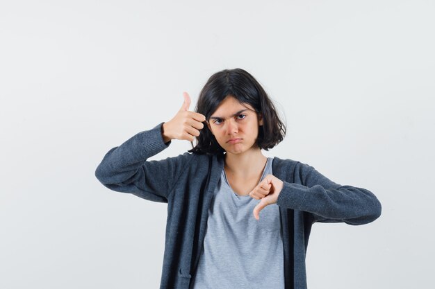 Ragazza che mostra il pollice su e giù in t-shirt grigio chiaro e felpa con cappuccio con zip grigio scuro e sembra seria