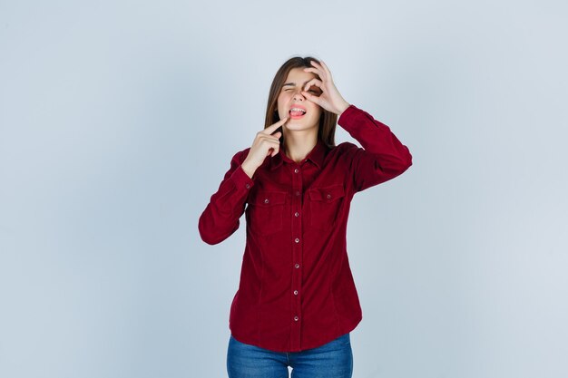 ragazza che mostra il segno ok sull'occhio, indicando la lingua in una camicia casual e guardando pensierosa