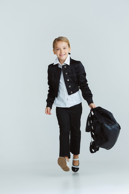 Ragazza che prepara per la scuola dopo una lunga pausa estiva. Di nuovo a scuola. Piccolo modello caucasico femminile che posa nell'uniforme della scuola con lo zaino sulla parete bianca. Infanzia, educazione, concetto di vacanze.