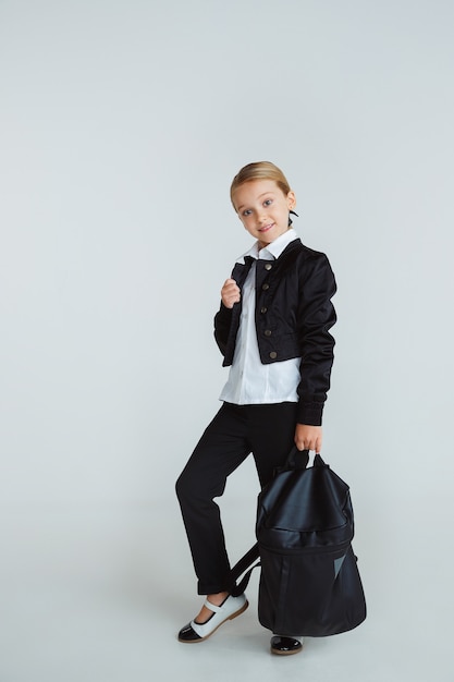 Ragazza che prepara per la scuola dopo una lunga pausa estiva. Di nuovo a scuola. Piccolo modello caucasico femminile che posa nell'uniforme della scuola con lo zaino sulla parete bianca. Infanzia, educazione, concetto di vacanze.