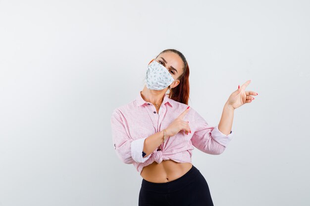 Ragazza che punta a destra con le dita indice in camicetta rosa, pantaloni neri, maschera e dall'aspetto seducente. vista frontale.