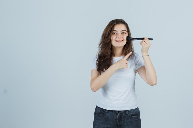 Ragazza che punta il pennello per il trucco in t-shirt, jeans e sembra carino vista frontale.