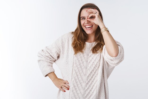 Ragazza che sbircia sorridente ampiamente godendosi le giornate ventose di primavera indossando un maglione alla moda sciolto che mostra il gesto ok ok guardando attraverso il cerchio tenendo tutto sotto controllo