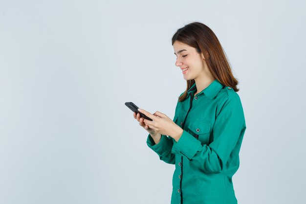 Ragazza che scrive messaggi sul telefono in camicetta verde, pantaloni neri e sembra allegro, vista frontale.