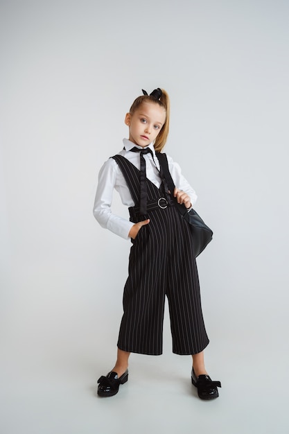 Ragazza che si prepara per la scuola dopo una lunga pausa estiva. Di nuovo a scuola. Piccola modella caucasica femminile in posa in uniforme scolastica con zaino su sfondo bianco. Infanzia, istruzione, concetto di vacanze.