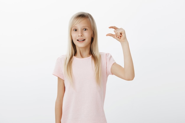 Ragazza che si sente ottimista ed eccitata, condividendo impressioni dopo aver visitato lo zoo. Figlia bionda carina in maglietta rosa, alzando la mano e modellando cosa piccola o minuscola con espressione ammirata sul muro grigio