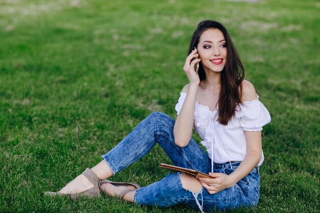 Ragazza che si siede in un parco parlare su un cellulare mentre si tiene un tablet