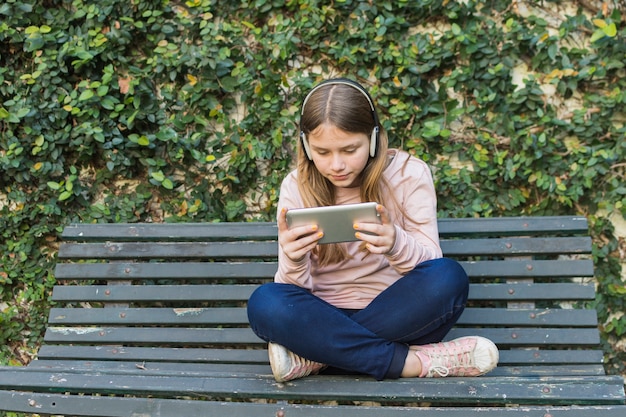 Ragazza che si siede sulla panchina che indossa la cuffia utilizzando il telefono cellulare nel parco