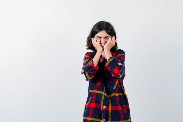 Ragazza che si tiene per mano sulle guance, fa una smorfia in camicia a quadri e sembra arrabbiata. vista frontale.