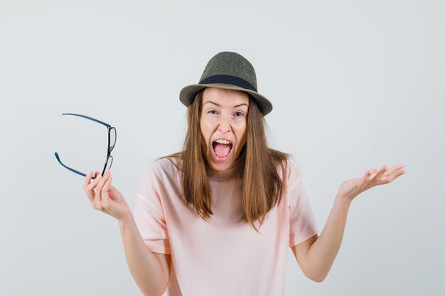 Ragazza che tiene gli occhiali mentre urla in maglietta rosa, cappello e sembra furioso. vista frontale.