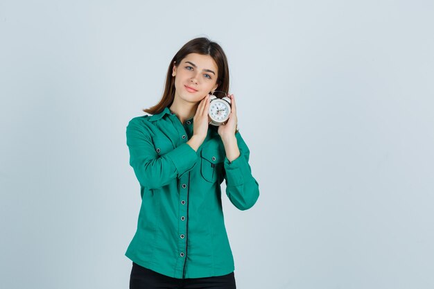 Ragazza che tiene l'orologio in entrambe le mani in camicetta verde, pantaloni neri e dall'aspetto sanguigno. vista frontale.