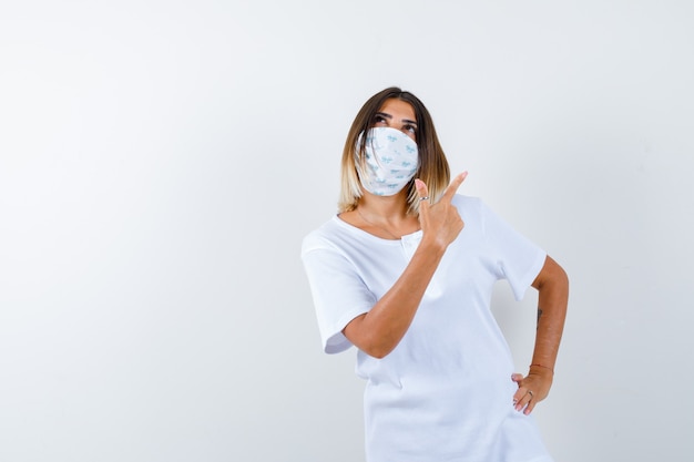 Ragazza che tiene la mano sulla vita, puntando a destra con il dito indice in maglietta bianca e maschera e guardando pensieroso. vista frontale.