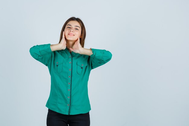 Ragazza che tiene le mani sul collo in camicetta verde, pantaloni neri e sembra carina. vista frontale.