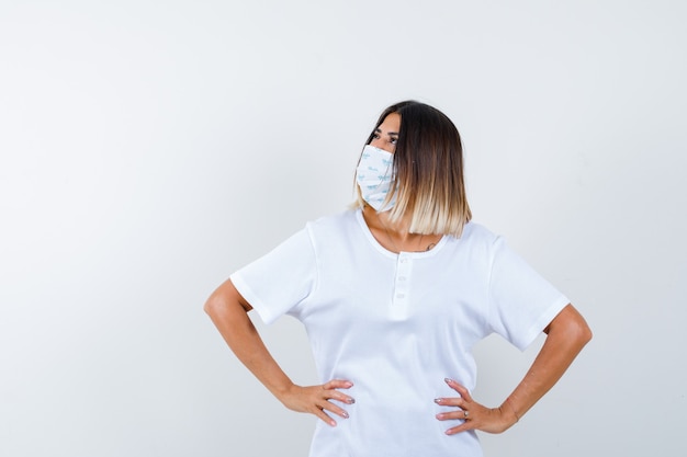 Ragazza che tiene le mani sulla vita, distogliendo lo sguardo in maglietta bianca e maschera e guardando concentrato. vista frontale.