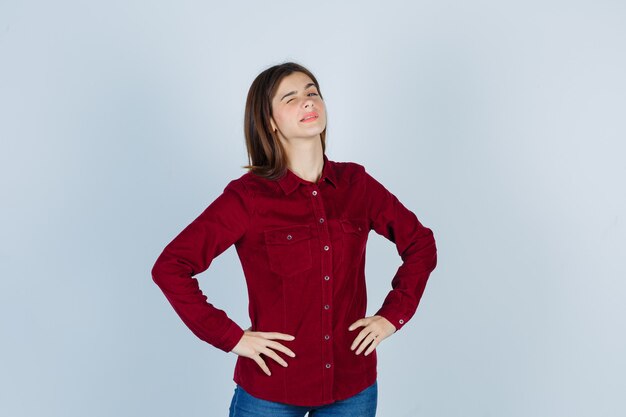 ragazza che tiene le mani sulla vita, sbatte le palpebre in camicia bordeaux e sembra sicura.