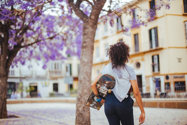 Ragazza che tiene skateboard che camminano sotto gli alberi in fiore