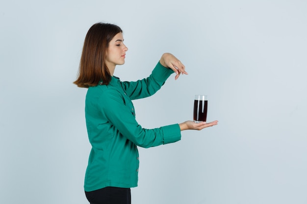 Ragazza che tiene un bicchiere di liquido nero, indicandolo con il dito indice in camicetta verde, pantaloni neri e guardando concentrato. vista frontale.