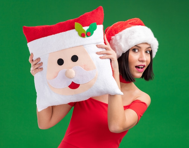 Ragazza colpita che porta il cappello della Santa che tiene il cuscino del Babbo Natale da dietro esso isolato sulla parete verde