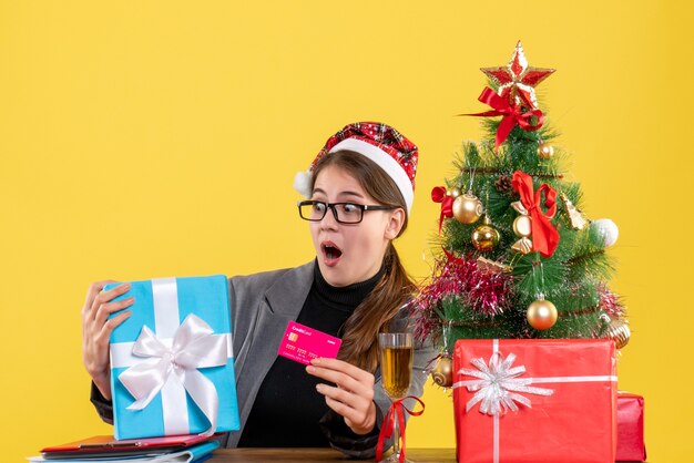 Ragazza con gli occhi spalancati di vista frontale con il cappello di natale che si siede al tavolo che tiene il cocktail dell'albero di Natale e dei regali della carta