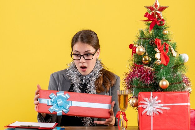 Ragazza con gli occhi spalancati vista frontale che si siede al tavolo guardando il suo regalo albero di Natale e regali cocktail