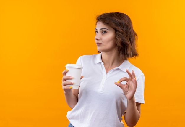 Ragazza con i capelli corti che indossa la camicia di polo bianca che tiene la tazza di caffè che sorride allegra mostrando segno giusto